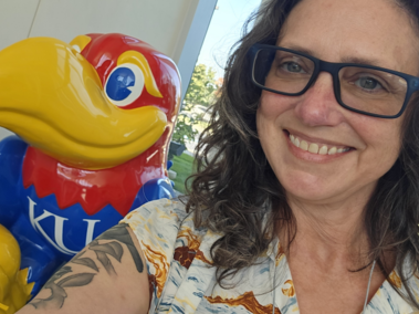 Birdie Ganz, an alum of the University of Kansas, smiling, wearing glasses and a beach-themed top, and standing in front of the KU Jayhawk on the Edwards Campus at KU.
