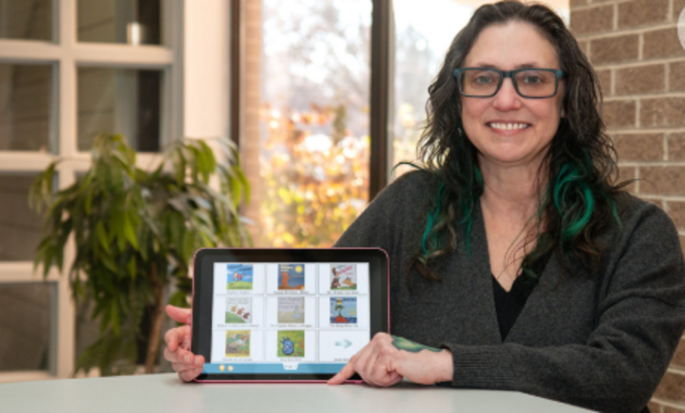 Birdie Ganz holding an AAC device and smiling, with brown and turquoise hair, glasses, and a gray sweater.